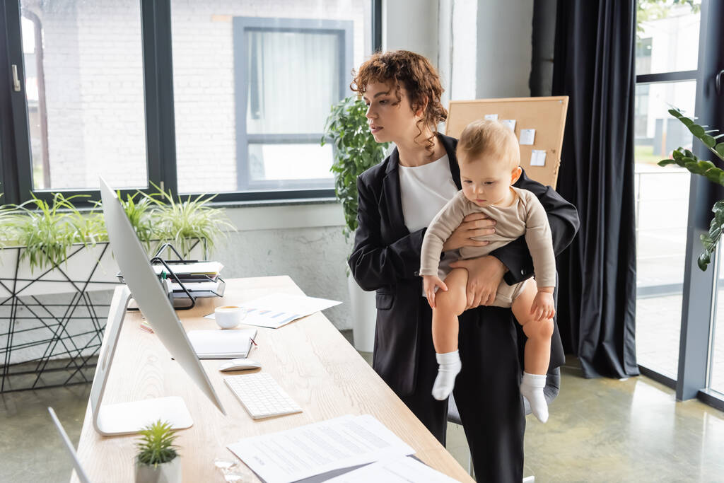 businesswoman in black suit looking at computer monitor while standing with little daughter at work desk in office - Foto, imagen