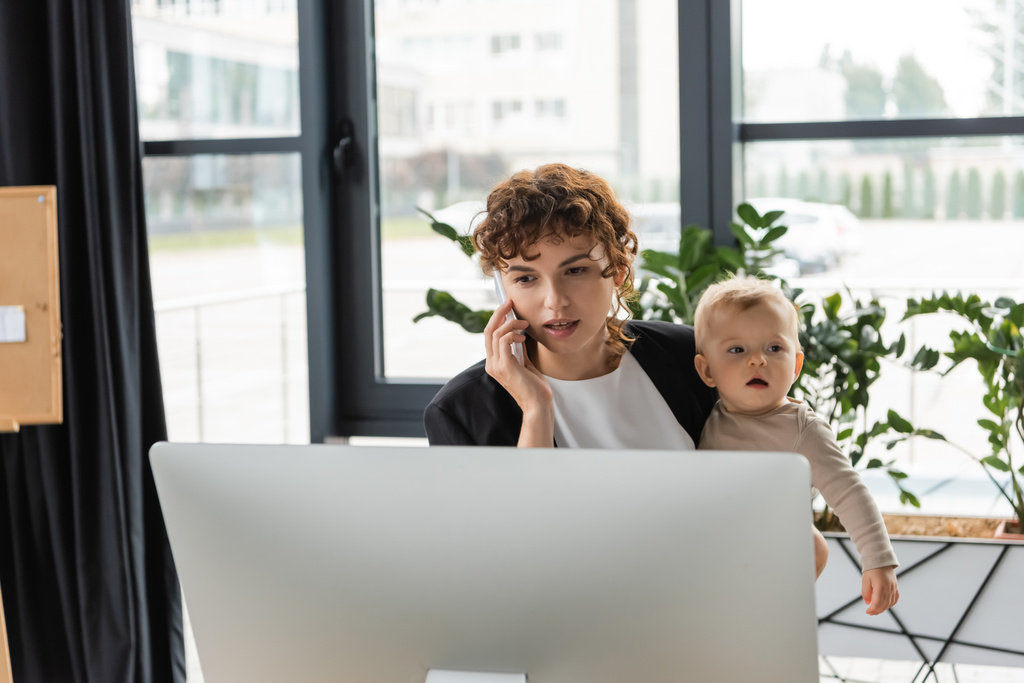 businesswoman calling on mobile phone while standing with toddler child near computer monitor - Foto, Bild