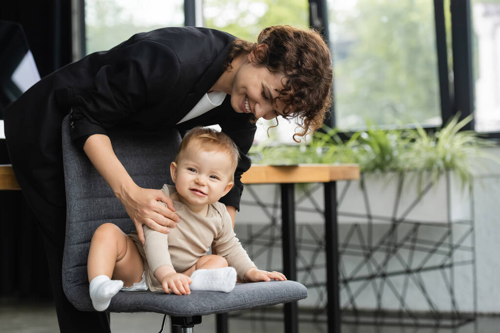 cheerful baby in romper sitting on office chair near smiling mother in formal wear - Photo, Image