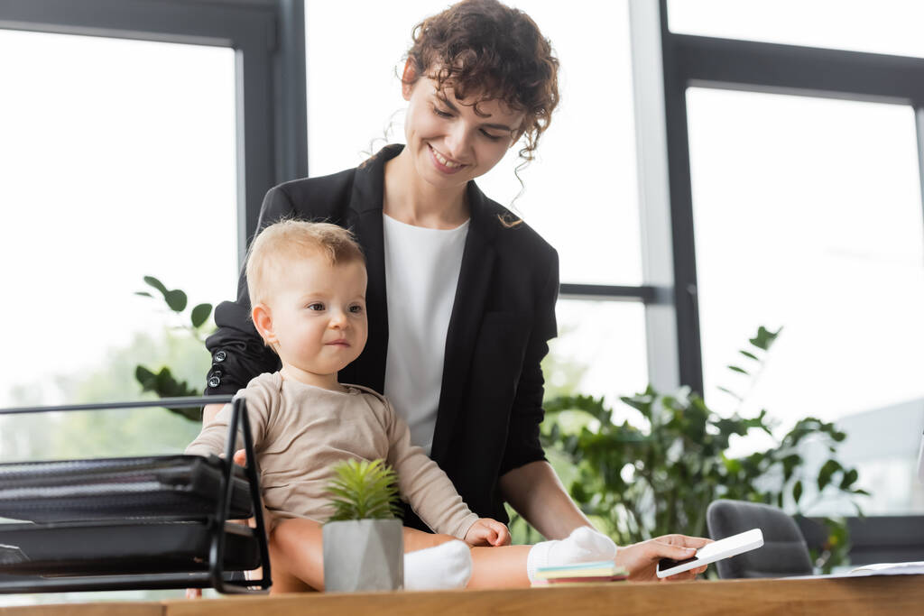 счастливая деловая женщина в черном пиджаке держит мобильный телефон рядом с дочкой малыша, сидящей на рабочем столе - Фото, изображение