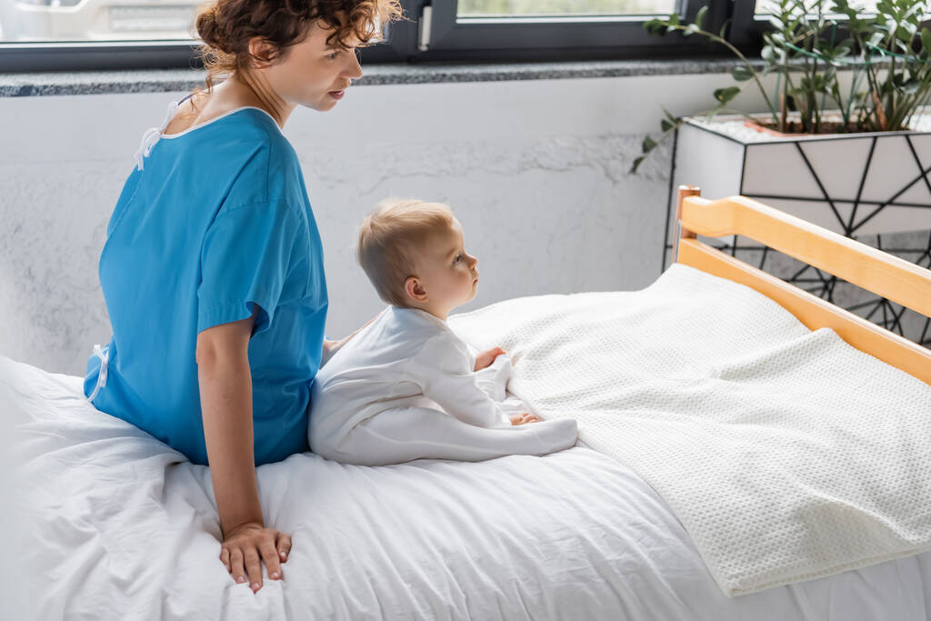 боковой вид женщины в больничном халате и малышки в комбинезоне, сидящей на кровати в клинике - Фото, изображение
