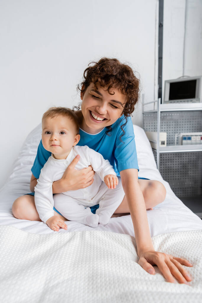 excited woman laughing with closed eyes and hugging baby on bed in hospital ward - Foto, Bild