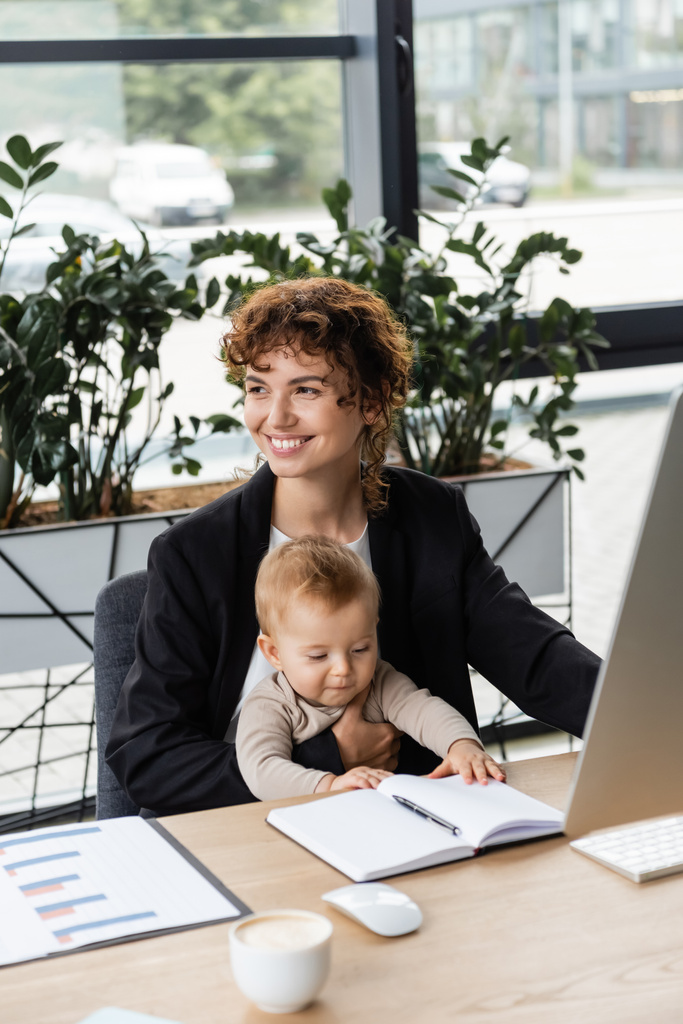 joyful businesswoman looking away while sitting with baby at work desk near blank notebook and blurred coffee cup - Foto, afbeelding