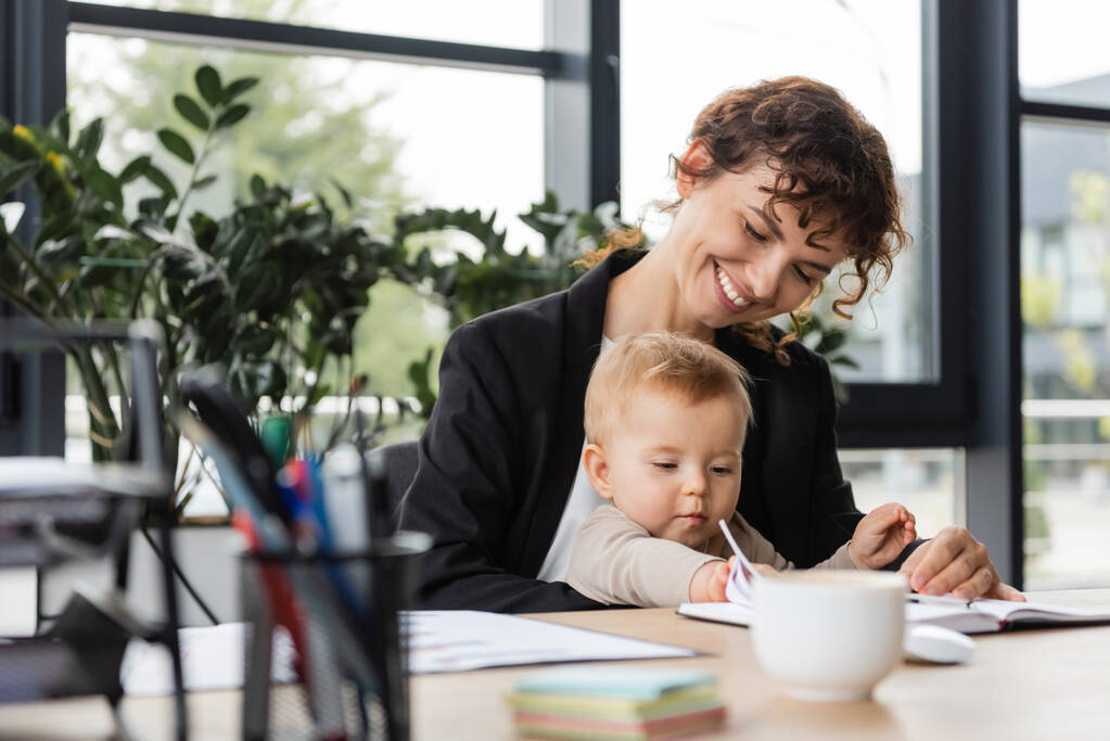 χαρούμενη επιχειρηματίας κάθεται στο χώρο εργασίας με την κόρη νήπιο κοντά σημειωματάριο και θολή φλιτζάνι καφέ - Φωτογραφία, εικόνα