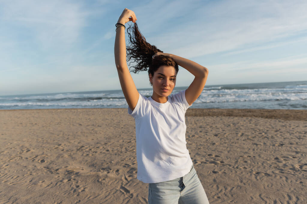 brunette woman in white t-shirt adjusting curly hair on sandy beach near sea  - Fotografie, Obrázek