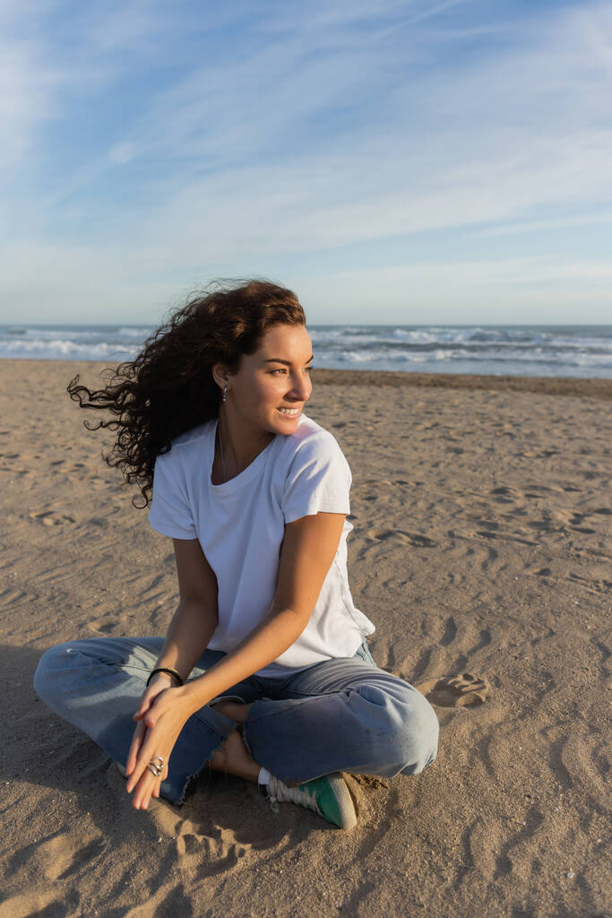 mujer positiva sentada en jeans azules y camiseta blanca sentada en la playa de arena en España  - Foto, imagen