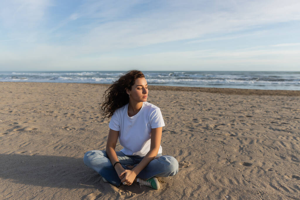 krullend brunette vrouw zitten in blauwe jeans en wit t-shirt zitten op zandstrand in Spanje  - Foto, afbeelding