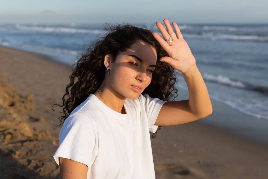 Porträt einer lockigen Frau im weißen T-Shirt, die ihr Gesicht vor der Sonne am Strand am Meer versteckt  - Foto, Bild