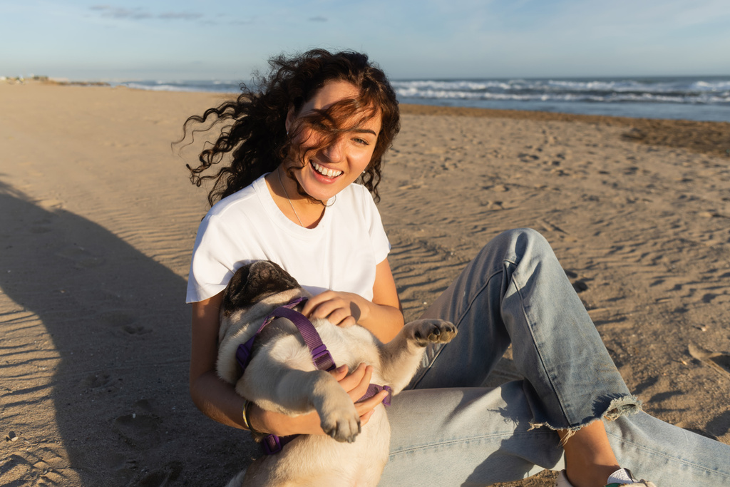 cheerful young woman with curly hair holding pug dog while sitting on sand near sea in Spain  - Photo, Image