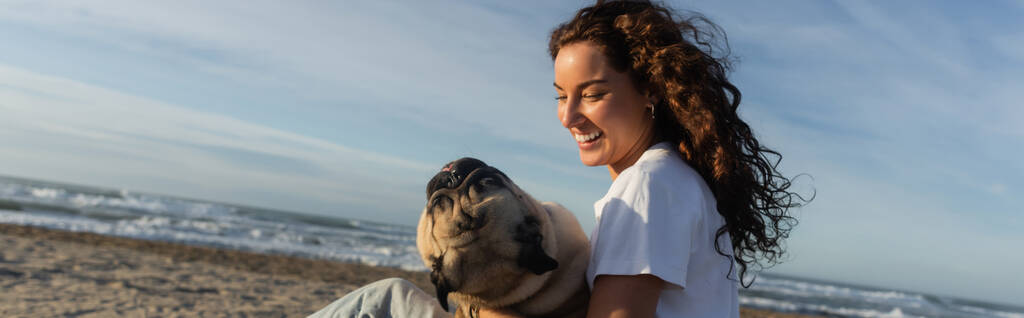 gelukkig jong vrouw met krullend haar vasthouden pug hond terwijl zitten op het strand in de buurt van de zee in Spanje, banner  - Foto, afbeelding