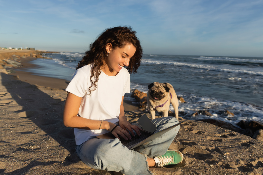 feliz joven freelancer con pelo rizado utilizando el ordenador portátil cerca de perro pug en la playa cerca del mar en España  - Foto, imagen