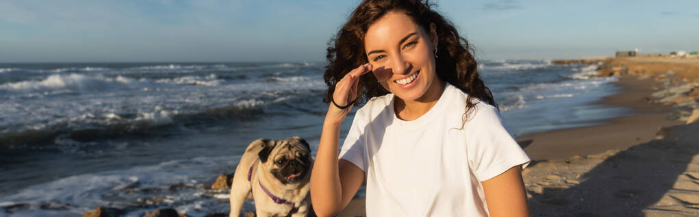 cheerful young woman covering cheek from sunshine near pug dog on beach near sea in Spain, banner  - Fotografie, Obrázek