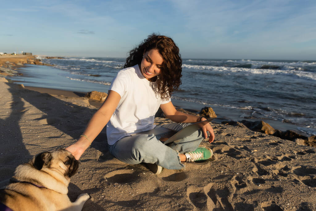 陽気なフリーランサーはノートパソコンと抱っこ犬とスペインの海の近くのビーチで座っている  - 写真・画像
