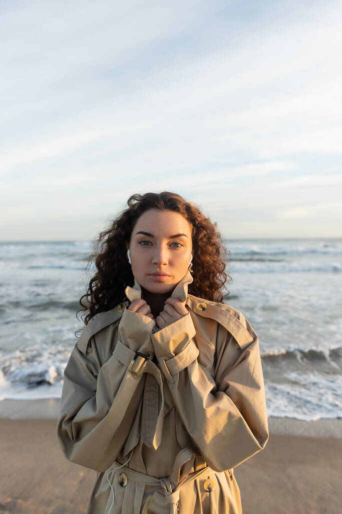curly young woman in wired earphones listening music while adjusting collar of beige trench coat near sea in Barcelona  - Foto, imagen