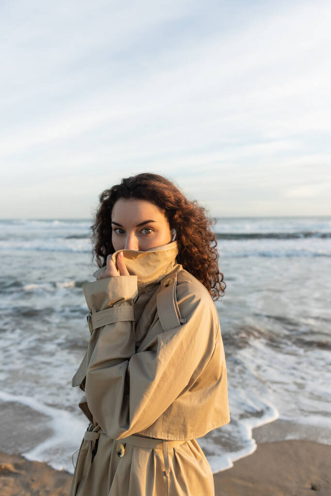 curly woman in wired earphones adjusting collar of beige trench coat and looking at camera near sea in Barcelona  - Photo, Image