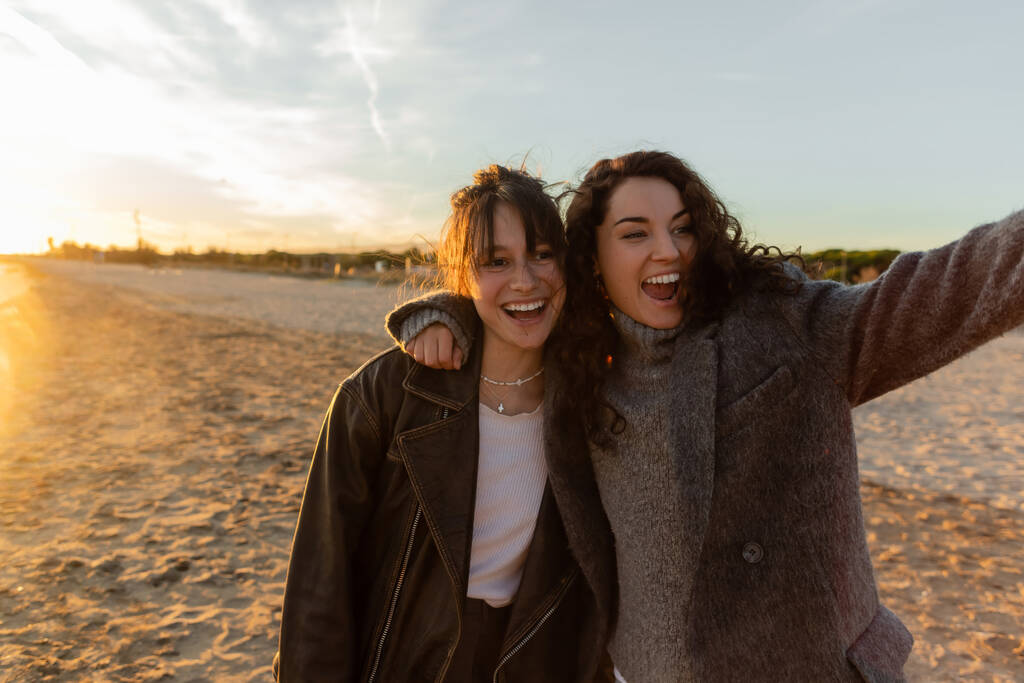 Cheerful curly woman hugging friend on beach during sunset  - Photo, Image