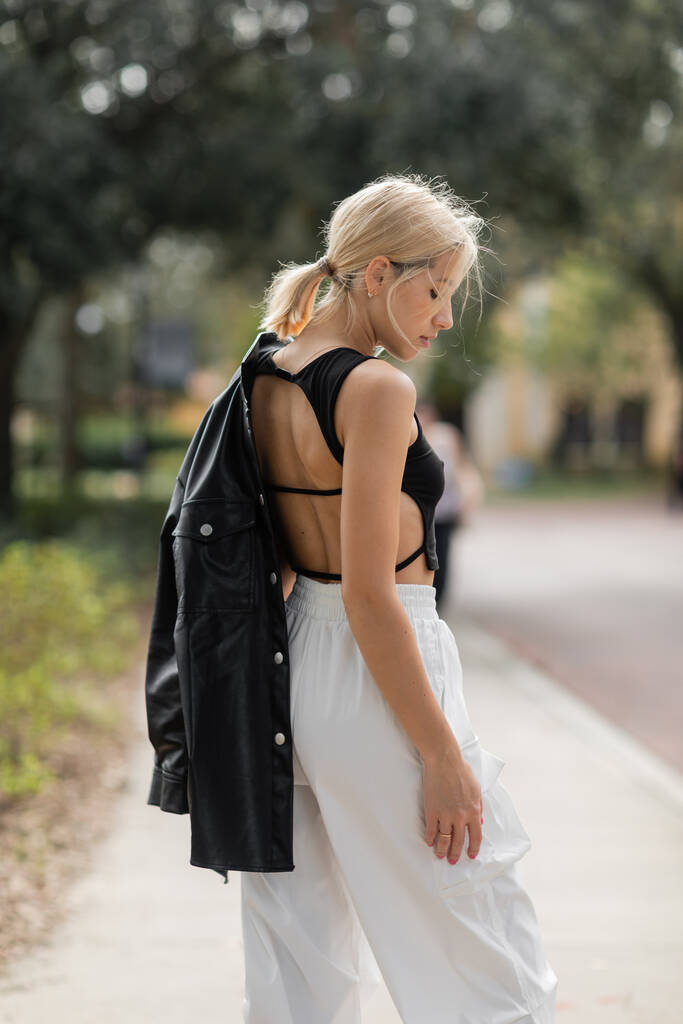 zijaanzicht van blonde vrouw in witte cargo broek staan met zwart lederen jas op straat in Miami   - Foto, afbeelding