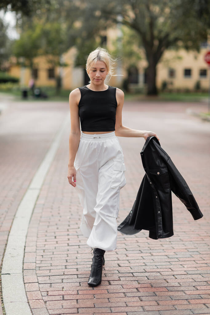 plná délka mladé ženy v bílém nákladu kalhoty a boty chůze s černou košilí bunda na ulici v Miami   - Fotografie, Obrázek
