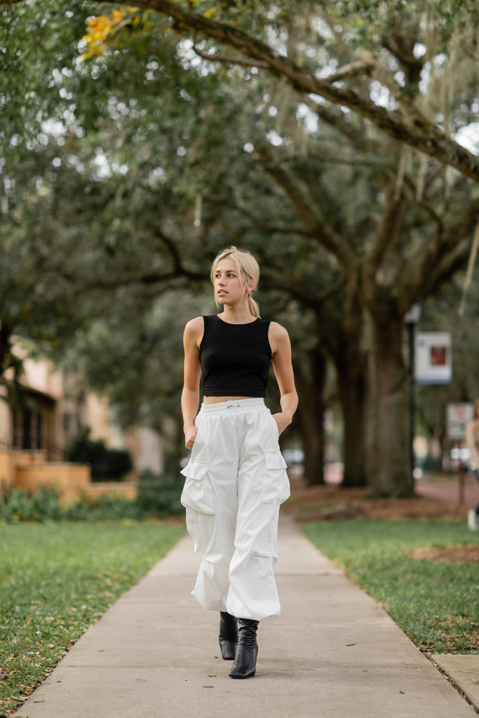 pełna długość blondynka kobieta w białych spodniach cargo i czarny top zbiornik chodzenie po zielonej ulicy w Miami - Zdjęcie, obraz