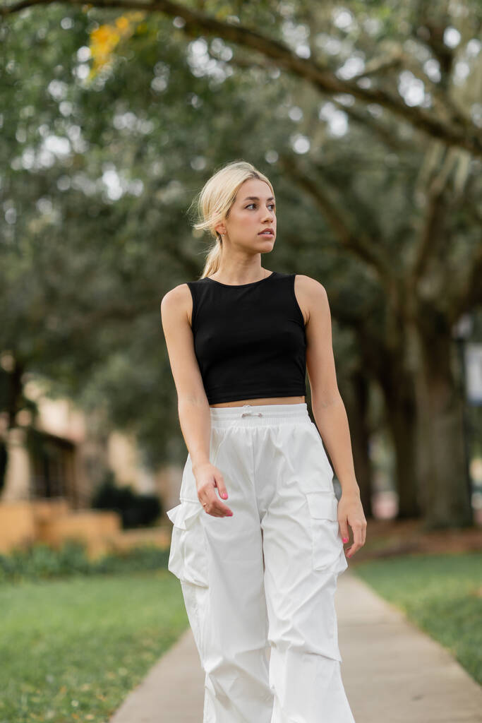 mladá blondýna žena v bílém nákladu kalhoty a černá tank top chůze na zelené ulici v Miami - Fotografie, Obrázek