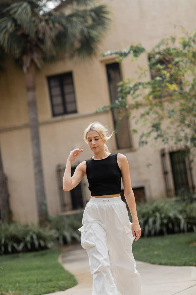 mujer joven en pantalones de carga blancos y camiseta sin mangas de moda caminando cerca de la casa en Miami - Foto, imagen