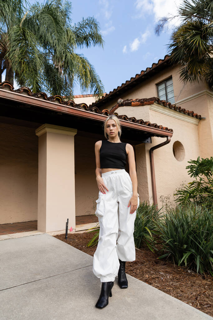 longitud completa de la mujer joven en ropa de moda y botas de pie cerca de la casa moderna y plantas verdes en Miami  - Foto, imagen