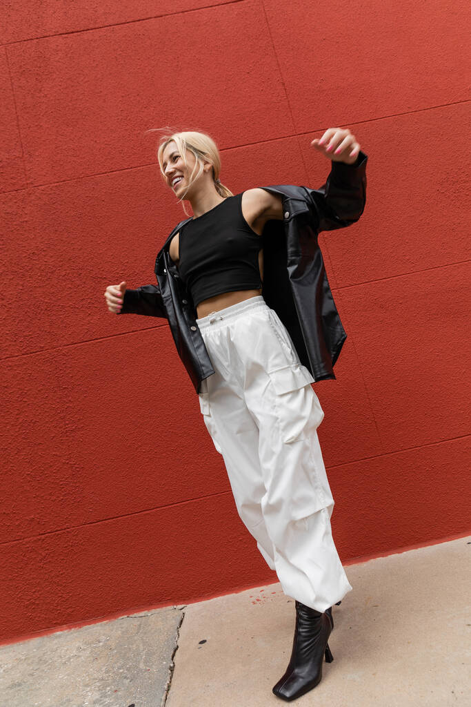 piena lunghezza di donna allegra in pantaloni cargo e giacca di camicia di pelle in piedi vicino al muro rosso a Miami  - Foto, immagini