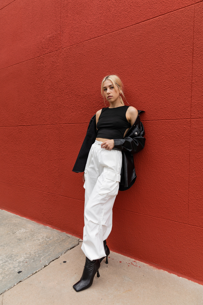 full length of blonde woman in cargo pants and leather shirt jacket standing near red wall in Miami  - Photo, Image