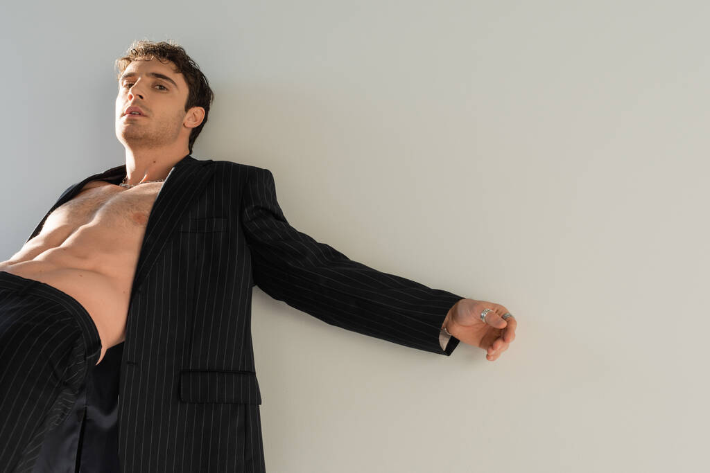 σέξι άντρας φορώντας μαύρο σακάκι σε γυμνό σώμα κοιτάζοντας κάμερα απομονωμένη σε γκρι - Φωτογραφία, εικόνα