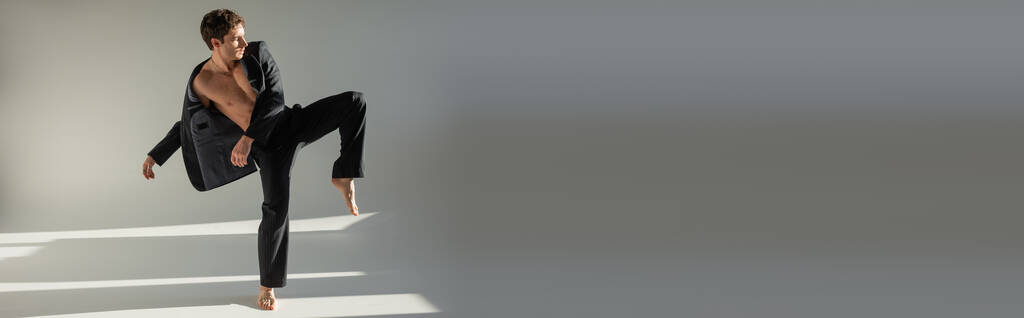 повнометражний модний босоніж чоловік в чорних штанях і блейзер на без сорочки тіло позує на одній нозі на сірому фоні, банер
 - Фото, зображення
