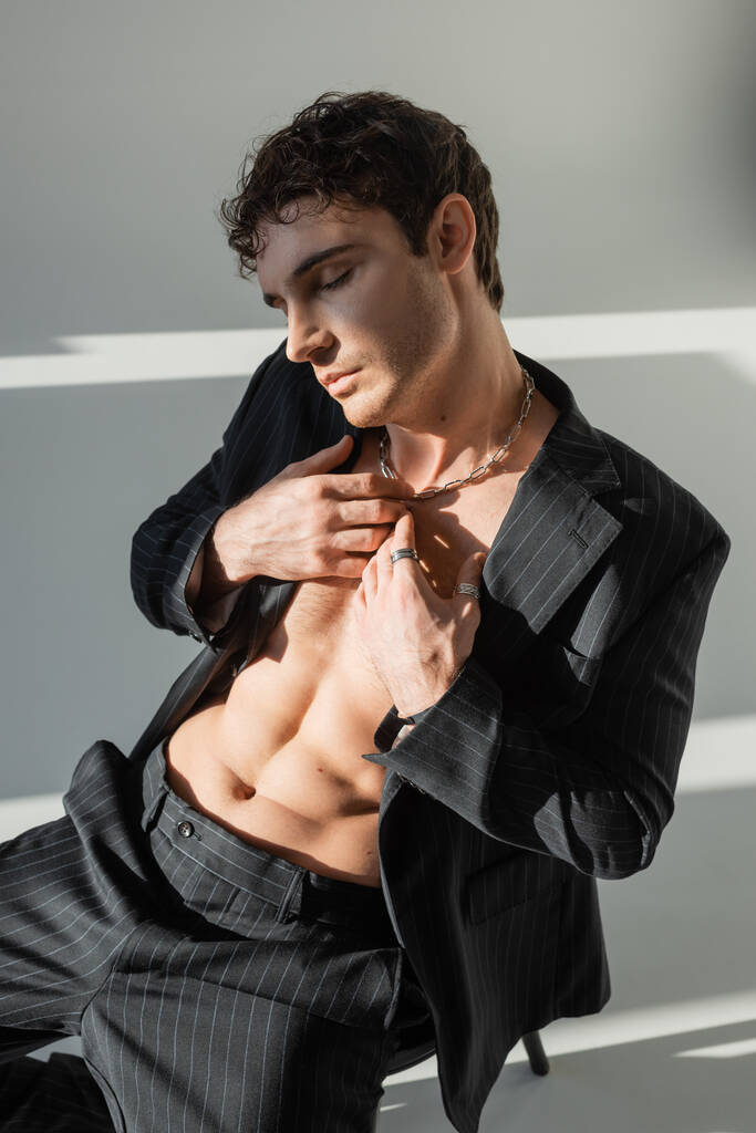man in silver necklace and black stylish blazer on shirtless torso sitting with closed eyes and hands near chest on grey background - Zdjęcie, obraz