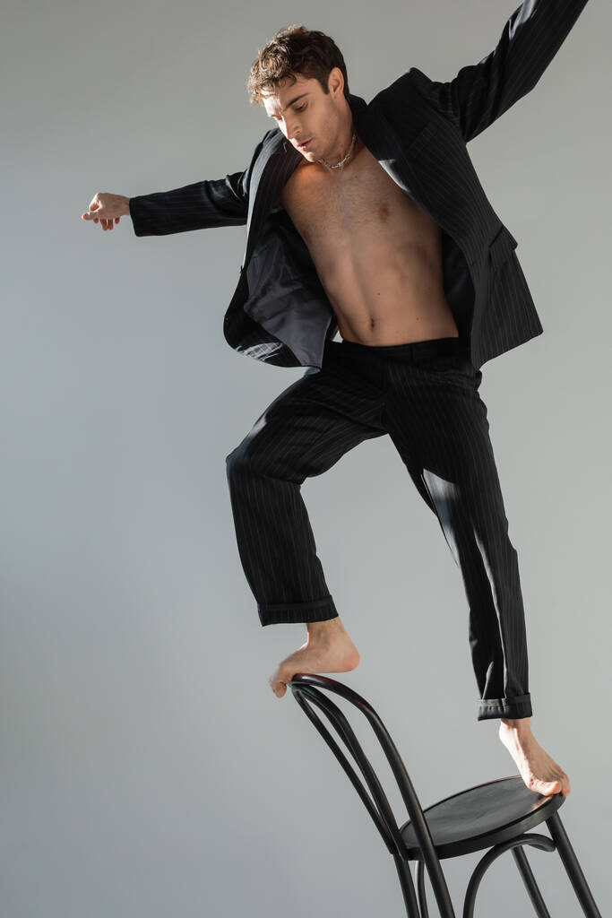 full length of barefoot man in black suit on shirtless body balancing on chair isolated on grey - Foto, Bild