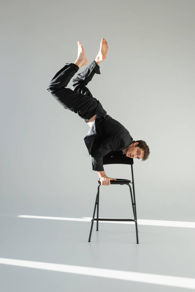 full length of barefoot man in black suit doing handstand on chair and looking at camera on grey background - Foto, imagen