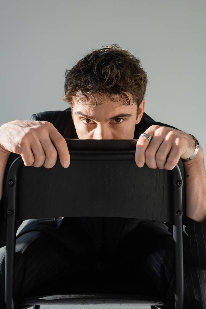 brunette man in black clothes hiding face behind chair back and looking at camera isolated on grey - Photo, image