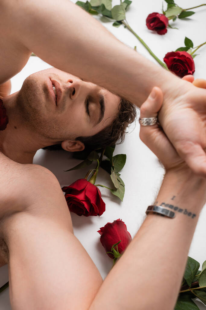 ylhäältä näkymä tatuoitu paidaton mies makaa kädet pään yläpuolella lähellä punaisia ruusuja valkoisella pohjalla - Valokuva, kuva