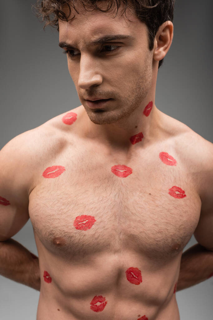 Мужчина без рубашки с красными поцелуями на мускулистом теле стоя с руками сзади на сером фоне - Фото, изображение