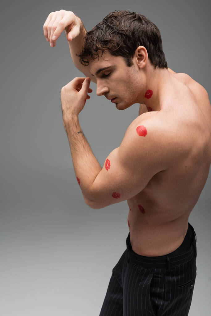 side view of brunette man with red lipstick marks on shirtless body posing on grey background - Photo, image