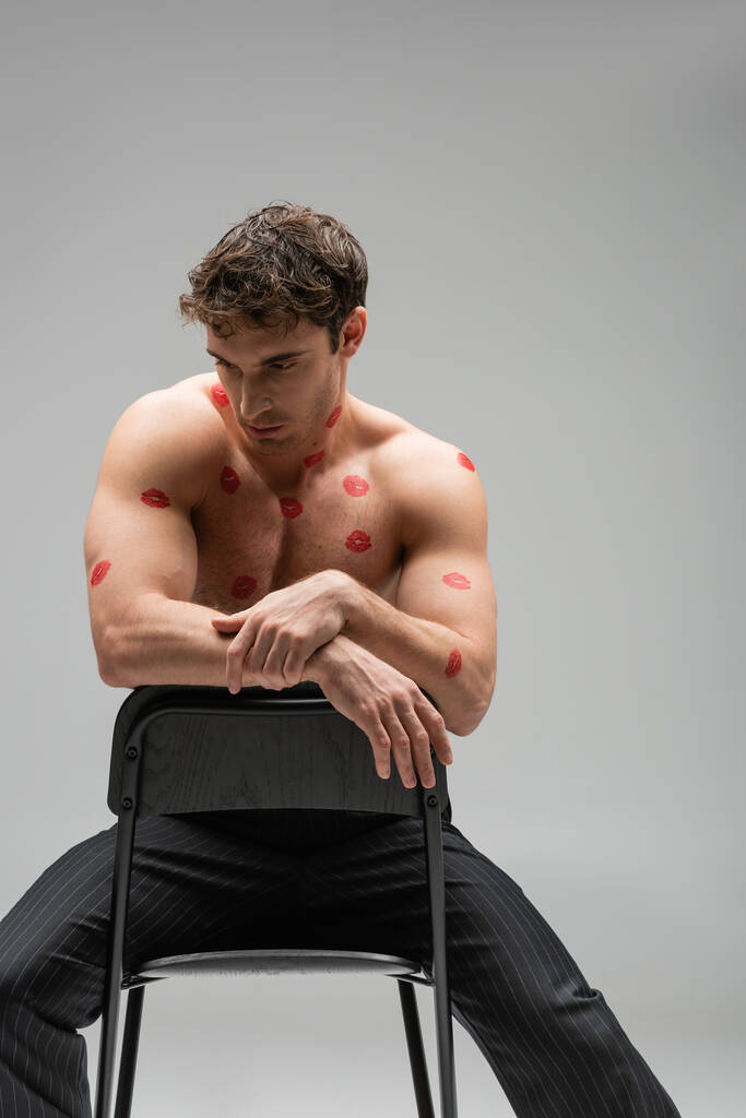 brunette muscular man with red kiss prints on body sitting on chair isolated on grey - Foto, afbeelding