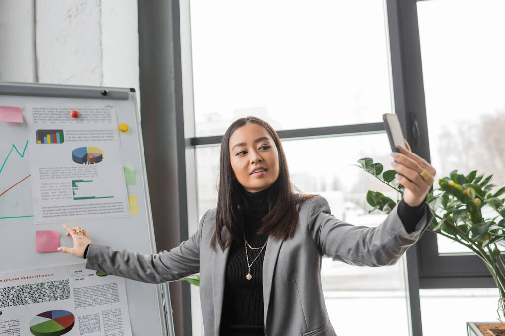 Asian designer pointing at flip chart during video call on smartphone in studio  - Fotografie, Obrázek