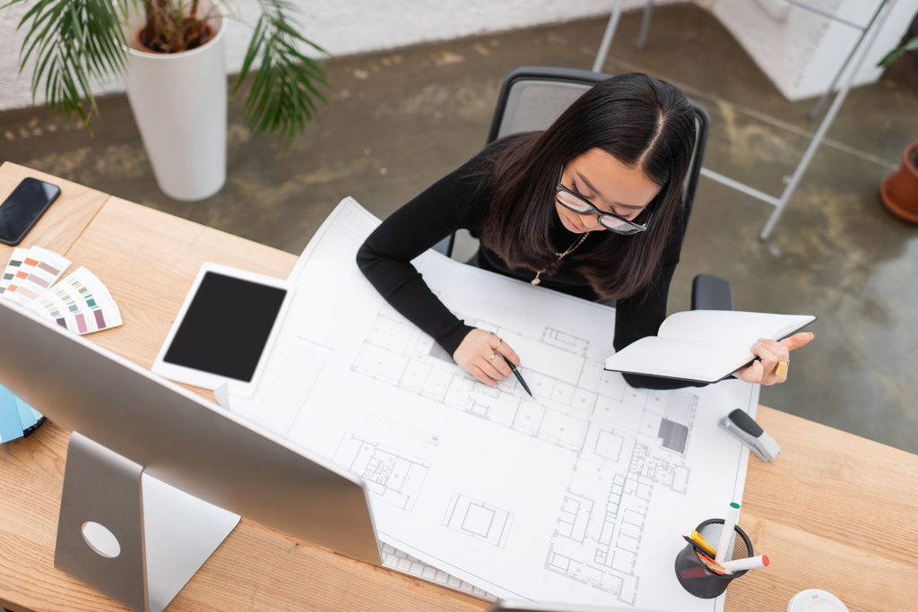 Вид сверху на азиатского дизайнера, работающего с чертежом и ноутбуком в офисе  - Фото, изображение