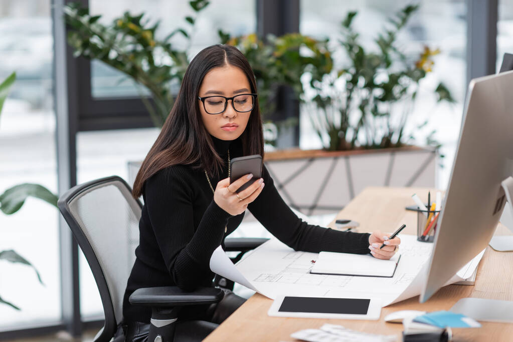 Brunette asian designer using smartphone near blueprint and notebook in office  - Photo, Image