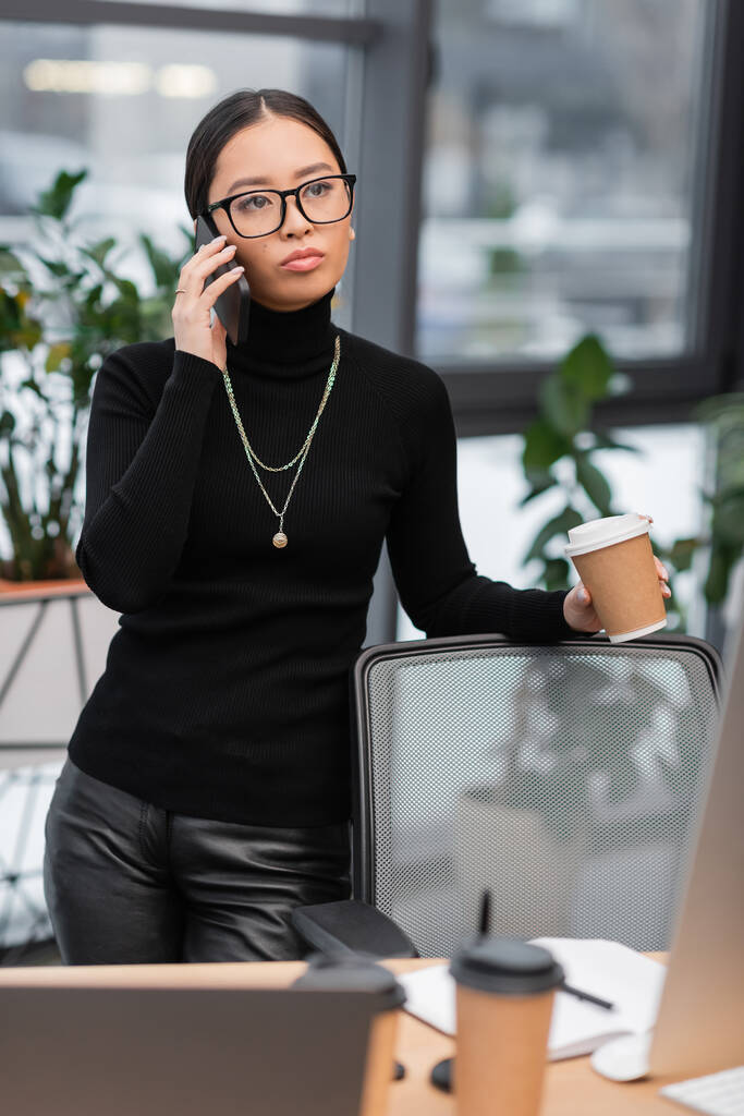 Asian interior designer talking on smartphone and holding coffee to go near devices in office  - Foto, imagen