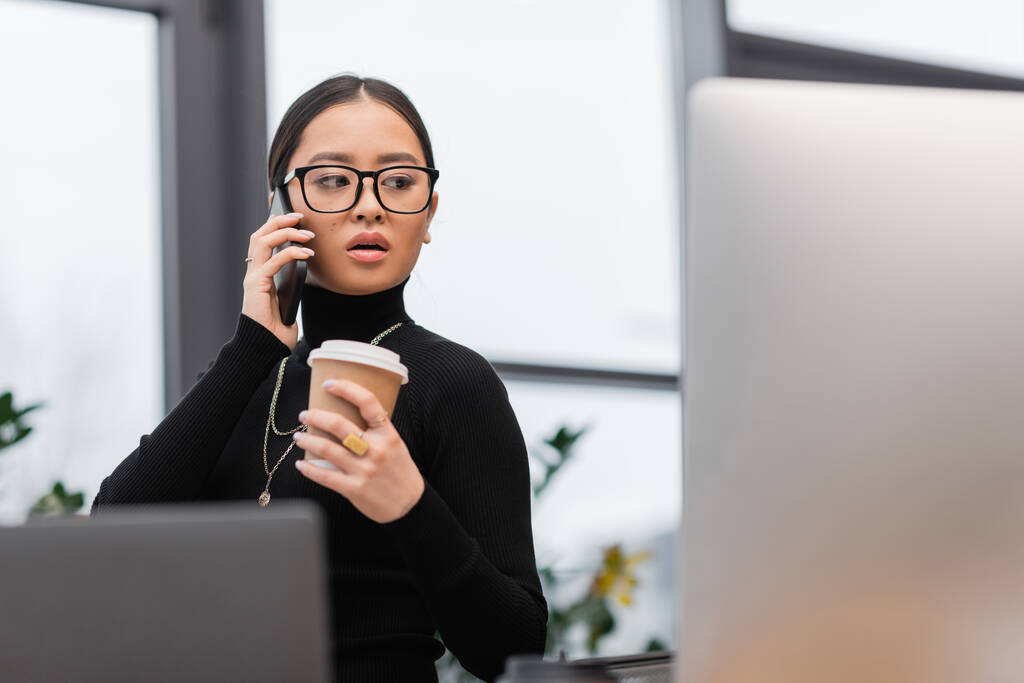 Азіатський дизайнер розмовляє на смартфоні і тримає каву біля комп'ютерів в офісі  - Фото, зображення