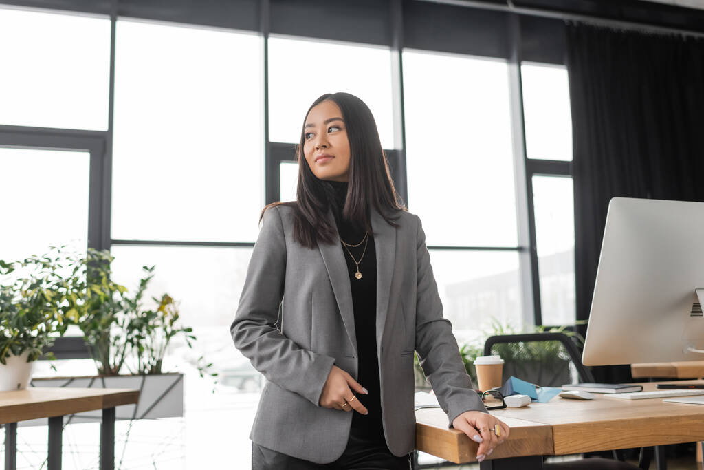 Brunette asian designer in jacket standing near working table in office  - Fotografie, Obrázek