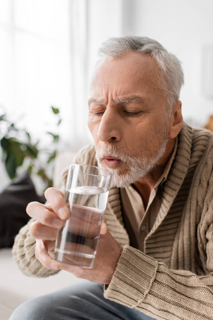 grey haired man with parkinsonism holding glass of water in trembling hands at home - Foto, afbeelding