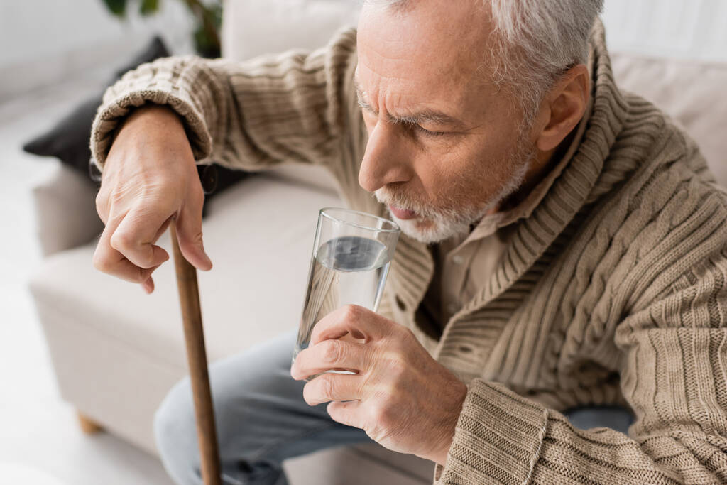 aged man with parkinson disease holding glass of water in trembling hand while sitting with walking cane at home - Foto, afbeelding