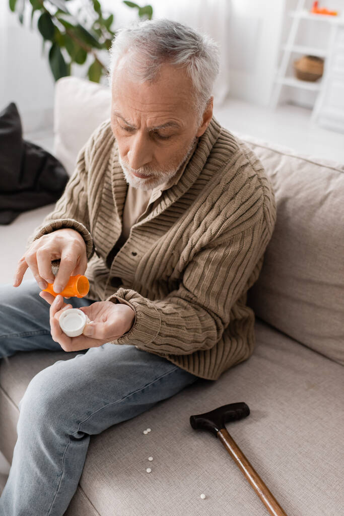 седовласый мужчина, страдающий паркинзонизмом и дрожью рук, держит контейнер с таблетками рядом с тростью для ходьбы на диване дома - Фото, изображение