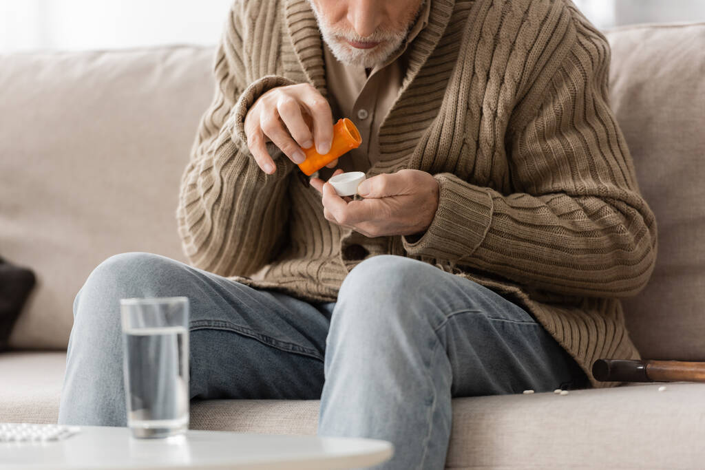partial view of aged man with parkinson syndrome sitting near blurred glass of water and holding pills container in trembling hands - Valokuva, kuva