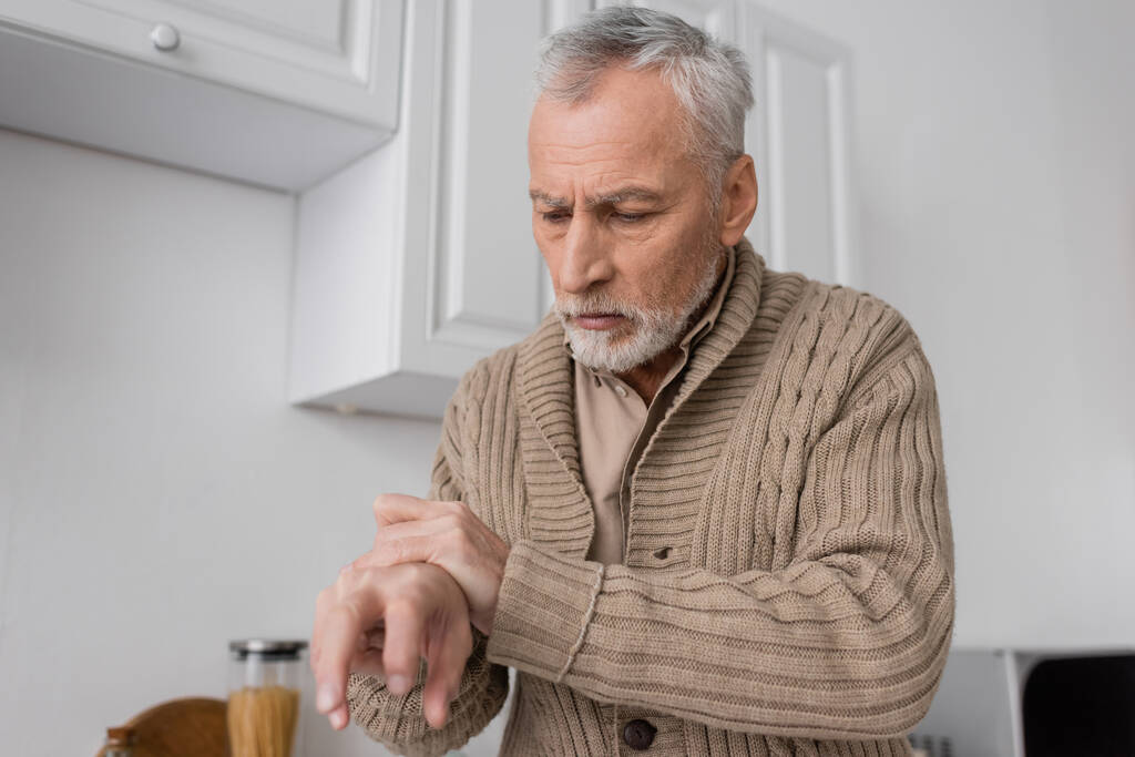 седой мужчина с болезнью Паркинсона смотрит на дрожащие руки на кухне - Фото, изображение