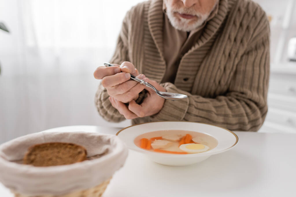 частичный вид пожилого человека с синдромом Паркинсона, сидящего с ложкой в дрожащих руках возле супа и хлеба на кухне - Фото, изображение
